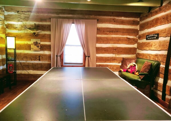 A ping pong table in a rustic cabin with wooden walls, a window with curtains, and a sign reading "The Gathering Place".