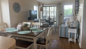 A dining area with a round table, white chairs, and a TV in the background. - Ocean-side condominium with pool and private beach access (Atlantic Beach)