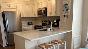 A modern kitchen with white cabinets, a stainless steel refrigerator, a microwave, a stove, and a sink. There are two white bar stools with woven seats. - Ocean-side condominium with pool and private beach access (Atlantic Beach)