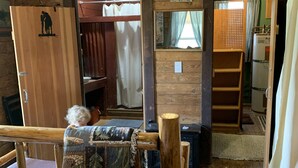 Interior rústico de una cabaña con una cama de madera, un banco y vistas a un área boscosa.