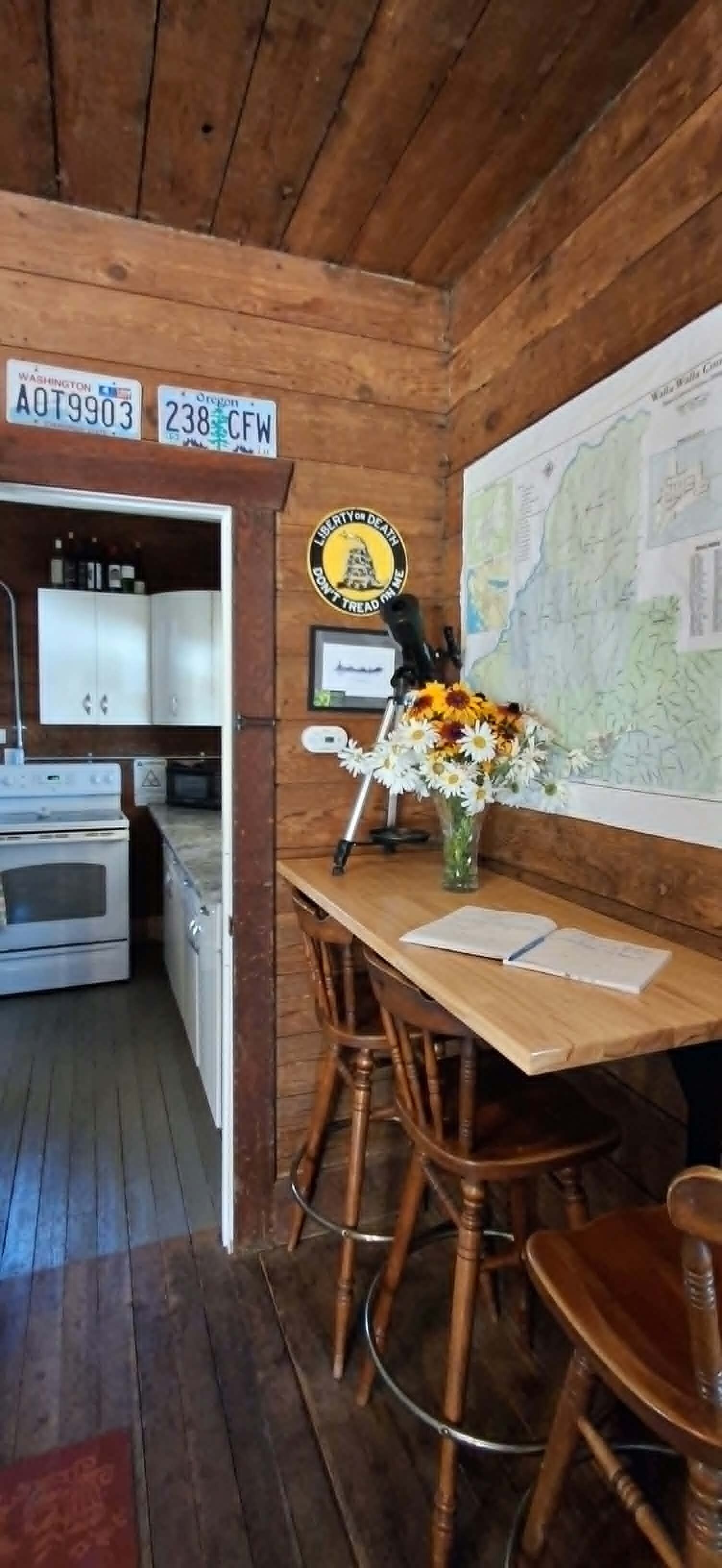 Un intérieur en bois comprenant une table à manger, des chaises, un vase avec des fleurs, une carte accrochée au mur et une cuisine visible à travers une porte ouverte.