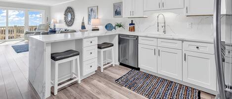 Una cocina moderna con armarios blancos, una isla central con taburetes, lavavajillas y vistas al exterior.