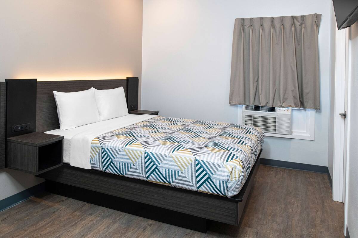 Un dormitorio con cama, mesitas de noche, cabecera, ventana con cortinas y un equipo de aire acondicionado.