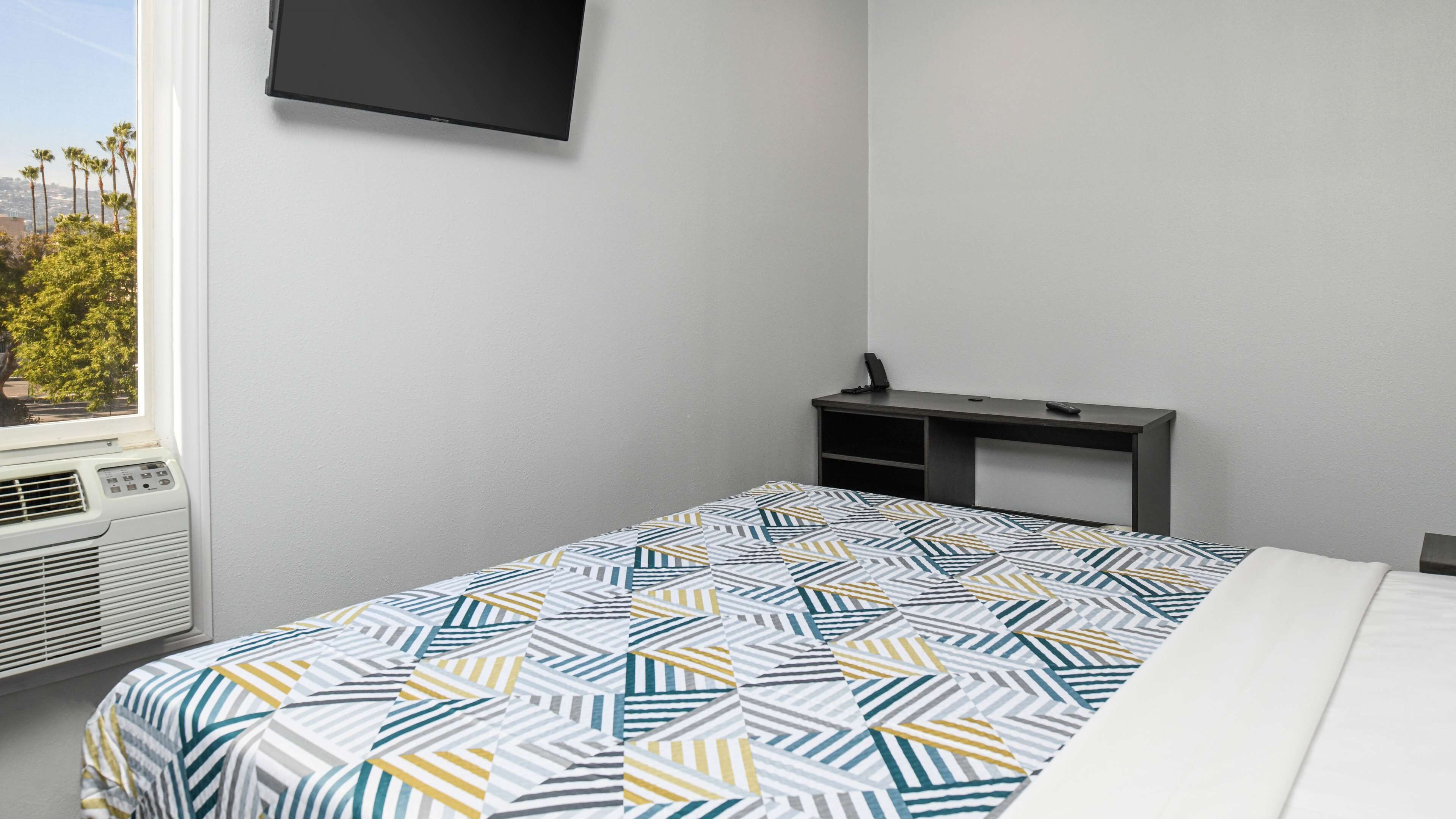 A hotel room with a bed, a TV mounted on the wall, a desk, and a window with a view of palm trees.