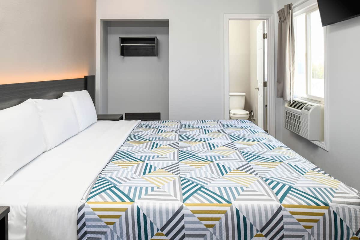 Una habitación de hotel con una cama, una ventana con cortinas, un aire acondicionado montado en la pared y una puerta que da a un baño.