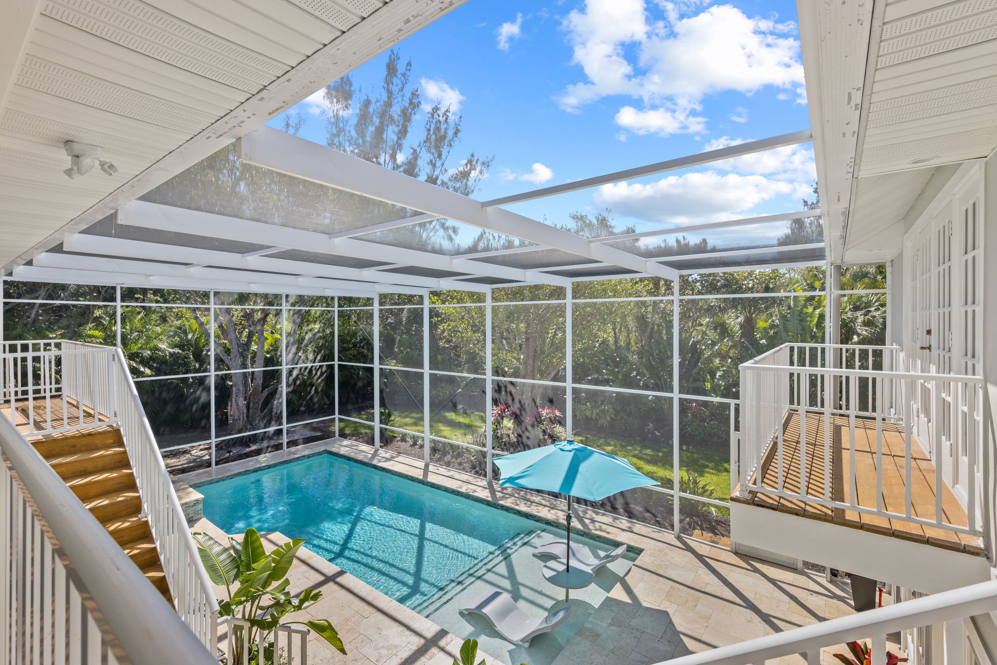 A covered outdoor swimming pool with a wooden deck, a blue umbrella, and a view of the surrounding greenery.