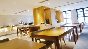A modern dining area with wooden tables and chairs, a kitchen area with appliances, and large windows allowing natural light. - Ji Hotel (Chengdu)