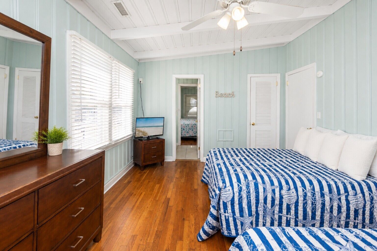 Une chambre avec une commode en bois, un lit avec une literie à rayures bleues et blanches, un ventilateur de plafond et une porte sur laquelle est inscrit le mot « Sœur ».