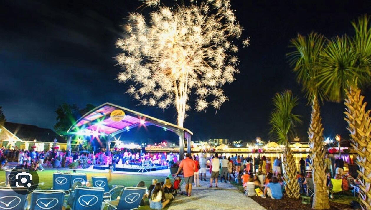 An outdoor event with a stage and audience, surrounded by palm trees and a large illuminated tree.