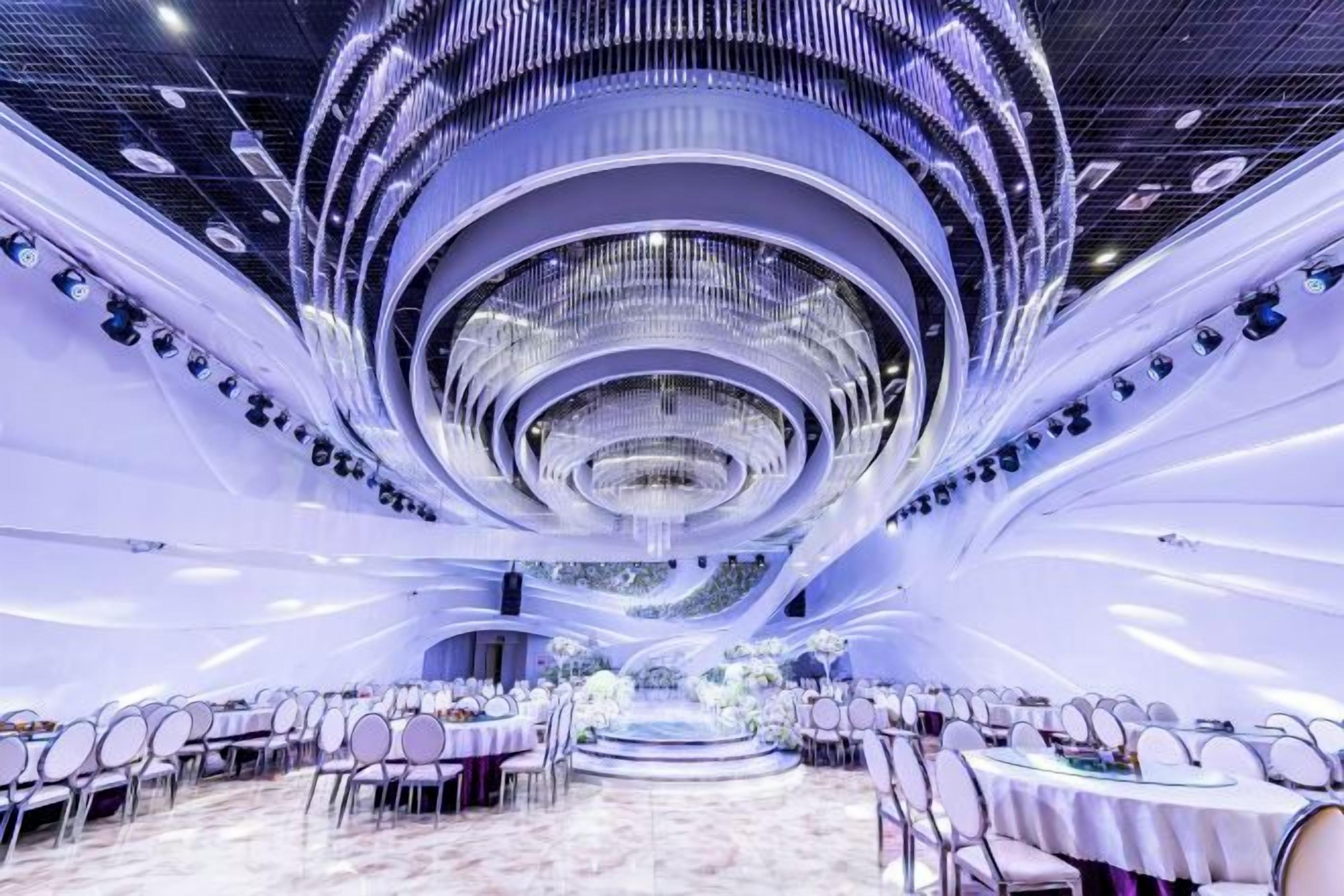 A spacious banquet hall with a central circular chandelier, multiple round tables, and a modern ceiling design.