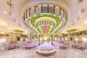 A spacious hall with a central circular table surrounded by pink chairs, a chandelier, and floral decorations.