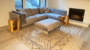 Un salon moderne avec un canapé d’angle, deux tabourets en bois, un tapis à motifs et une cheminée.