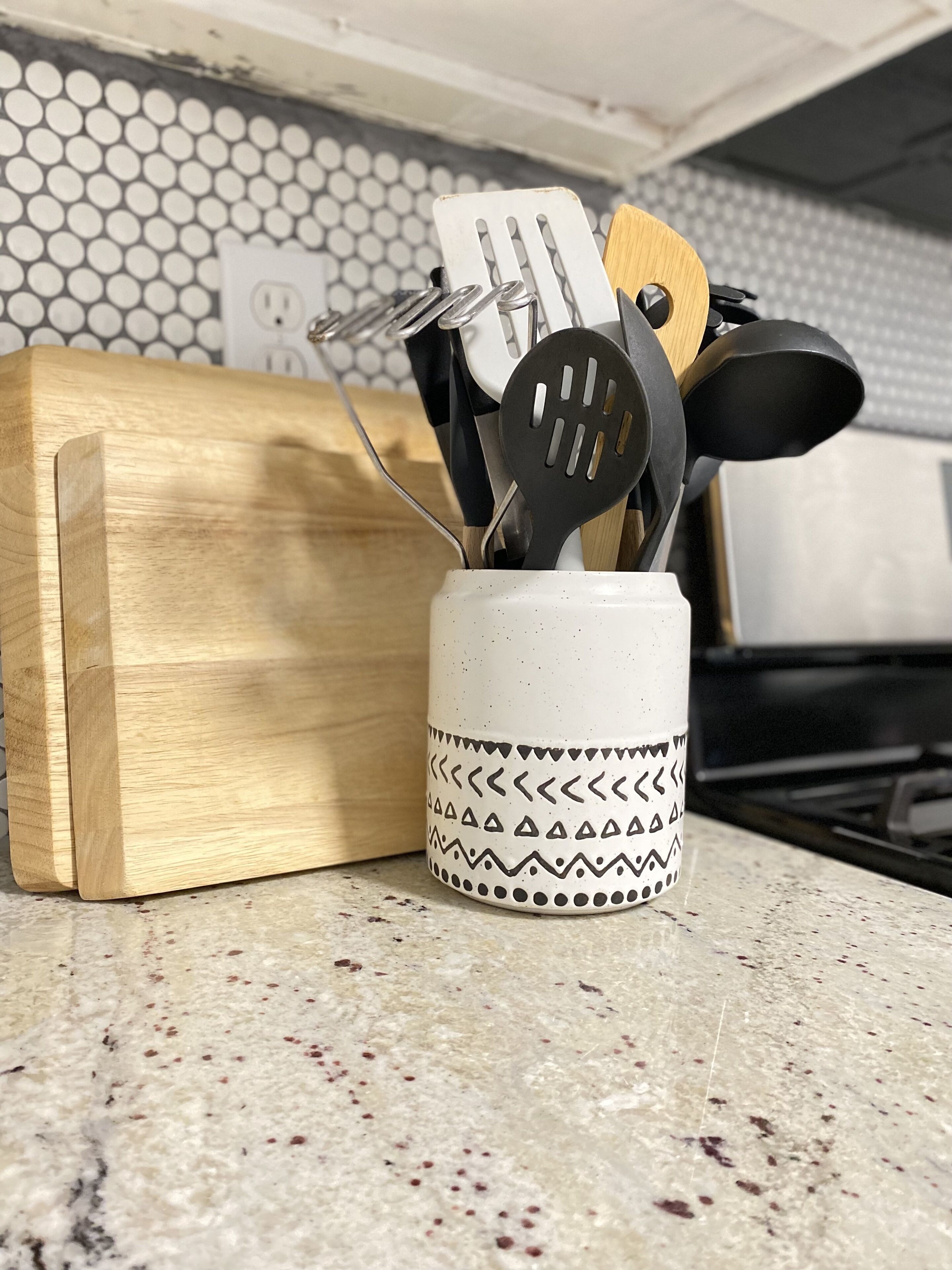 A kitchen counter with a wooden cutting board, a white mug with black patterns, and various kitchen utensils.
