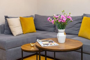Un sofá gris con cojines amarillos y blancos, un jarrón blanco con flores rosadas, una revista y una pequeña figurita de pájaro dorado sobre una mesa redonda de madera.
