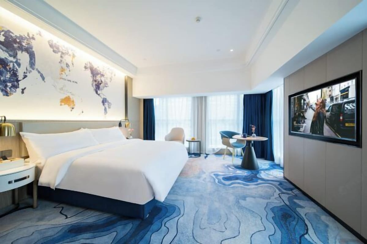 A hotel room with a large bed, a desk, a chair, a small table, a world map on the wall, and a TV.