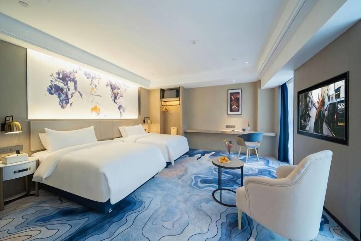 A hotel room with two beds, a round coffee table, a white armchair, a world map on the wall, and a large window.