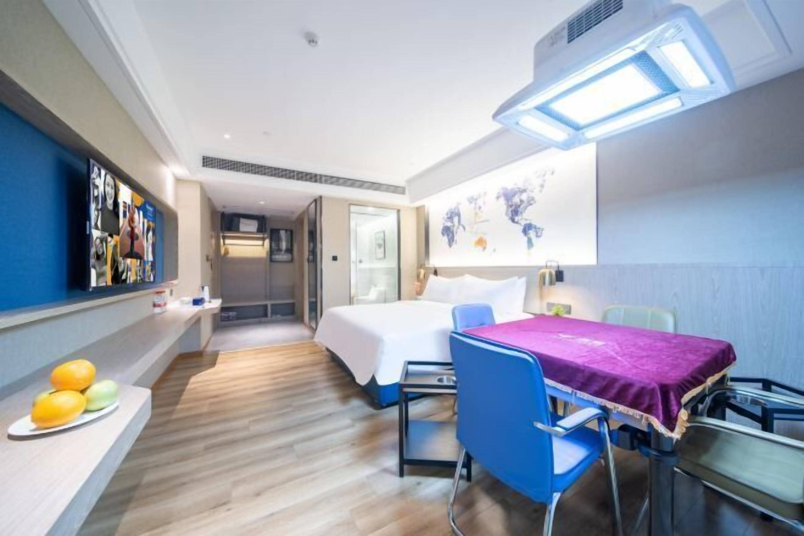 A hotel room with a bed, a desk with a purple tablecloth, a chair, a TV on the wall, and a fruit bowl on the side table.
