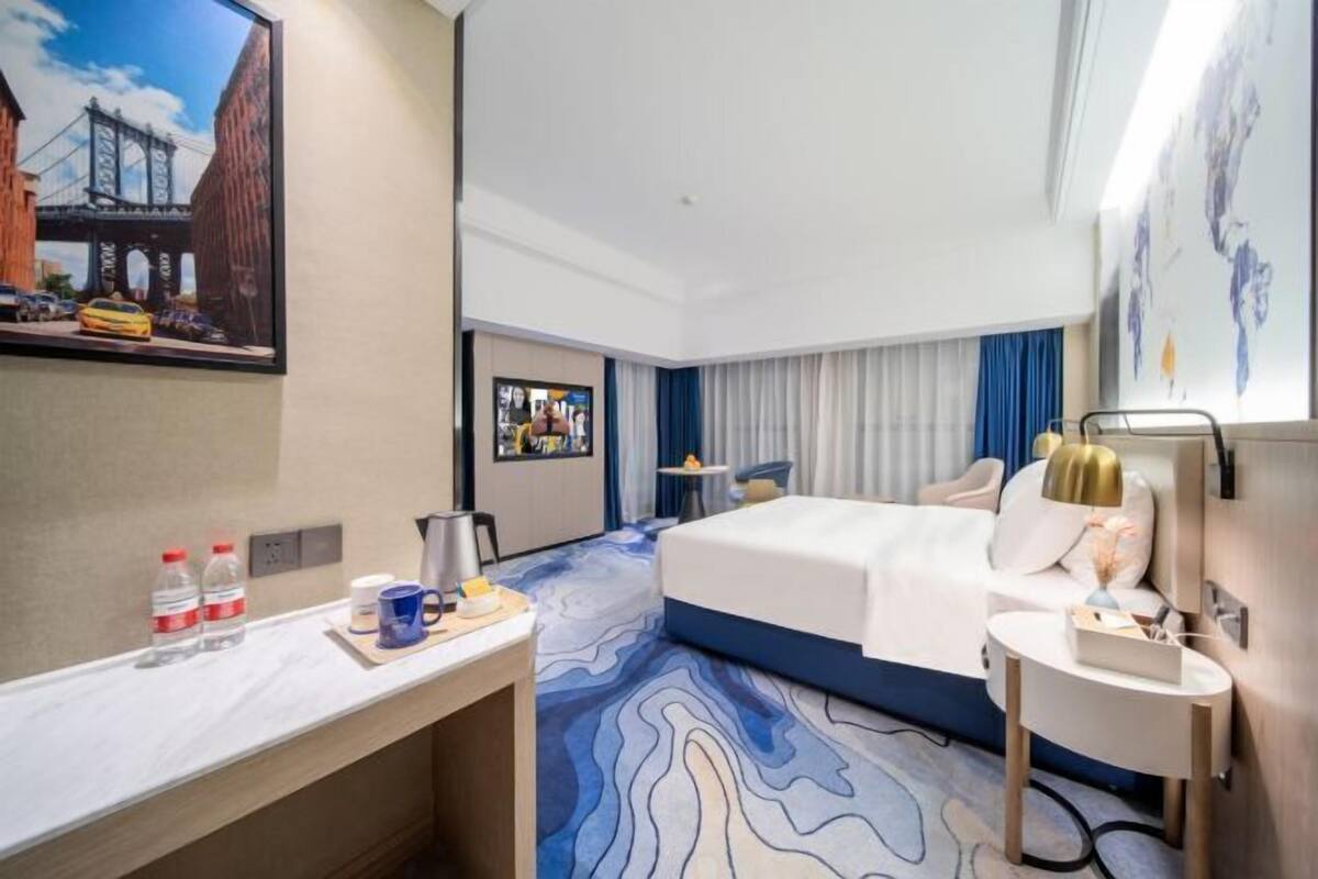 A hotel room with a bed, a desk, a chair, a small table, a TV, and a painting of a bridge.