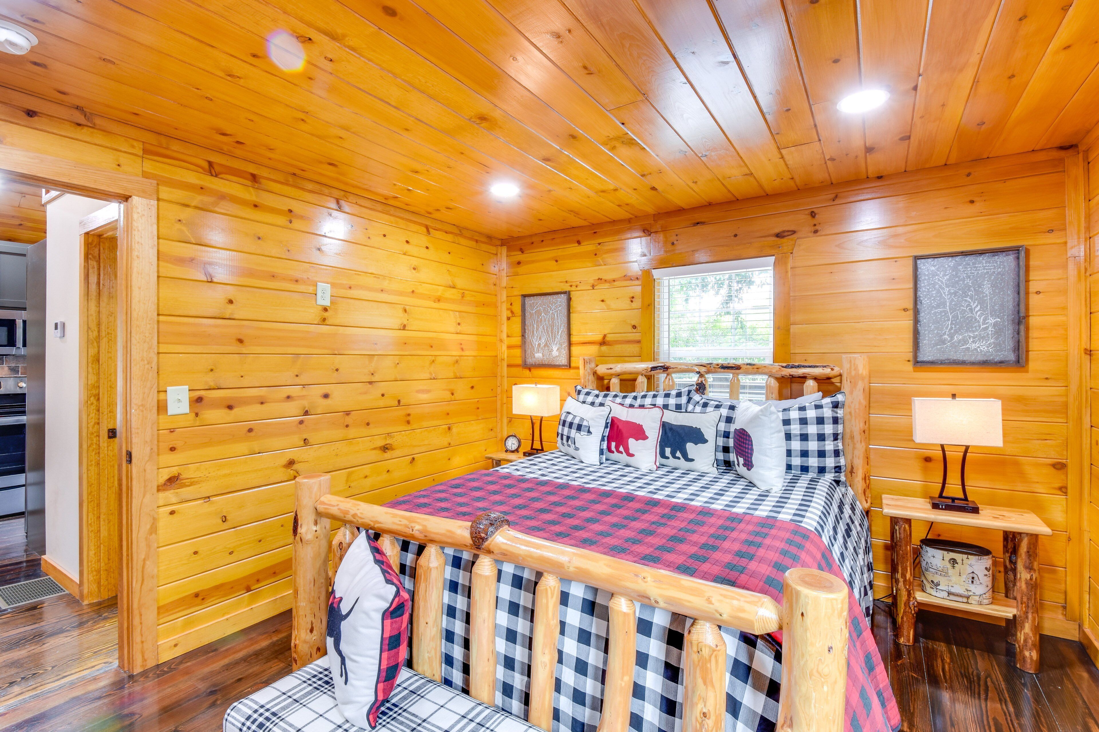 A log cabin bedroom with a wooden bed, checkered bedding, a nightstand with a lamp, and a window with a view of greenery.