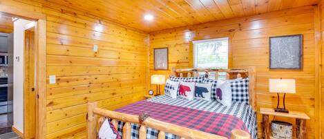 A log cabin bedroom with a wooden bed, checkered bedding, a nightstand with a lamp, and a window with a view of greenery.
