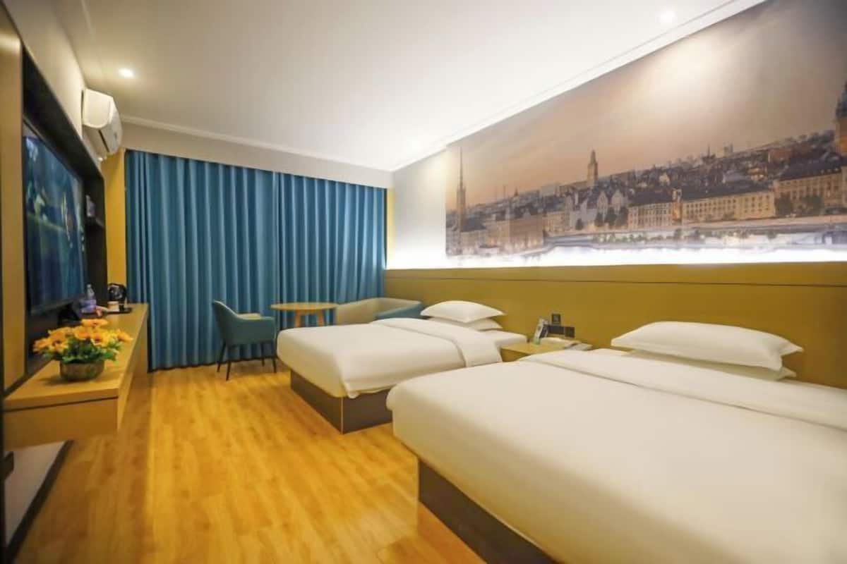 Habitación de hotel con dos camas, un escritorio con silla, un televisor y un cuadro de un paisaje urbano en la pared.