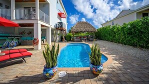 Una piscina con dos macetas, una estructura de techo de paja y un área de estar al aire libre cubierta con cojines rojos.