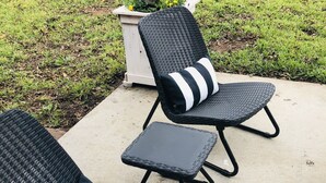 Dos sillones de mimbre al aire libre con cojines a rayas negras y blancas sobre un patio de concreto, rodeado de césped y un muro de ladrillo.