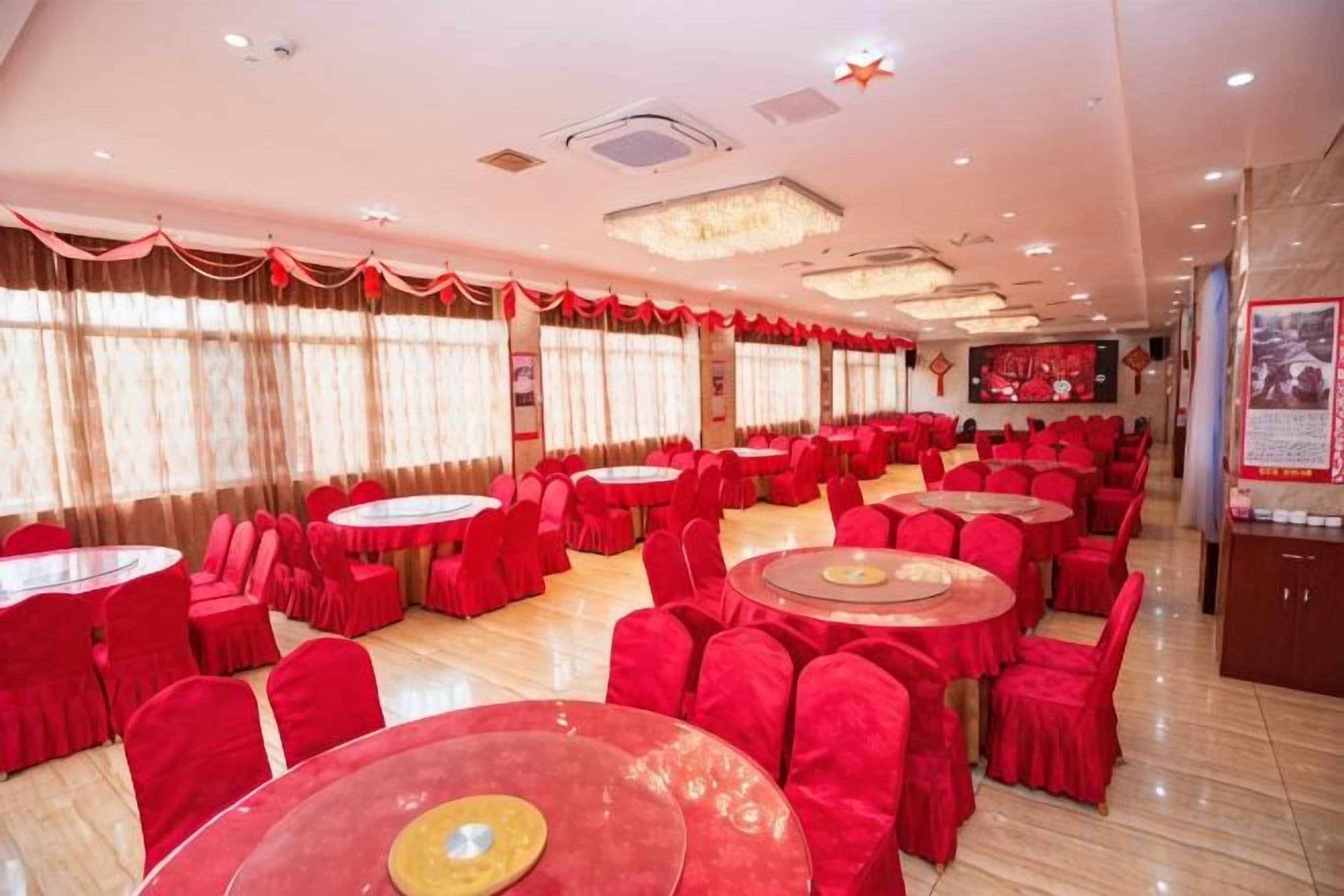 Un salón de banquetes con mesas redondas y sillas rojas, amplias ventanas con cortinas y una araña.