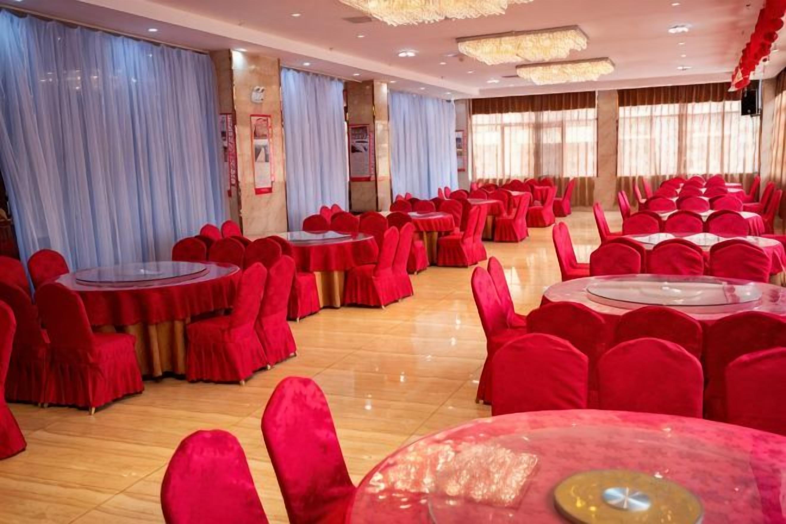 Un salón de banquetes con mesas redondas y sillas rojas, ventanas amplias y una araña.
