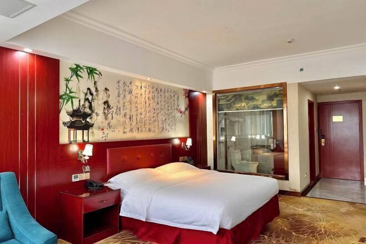 A hotel room with a large bed, a bedside table with a lamp, a chair, a wall with calligraphy, and a bathroom visible through a glass door.