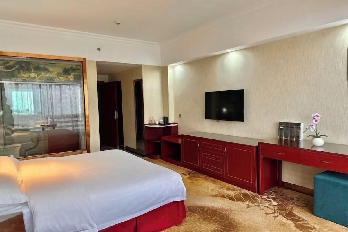 A hotel room with a large bed, a desk with a flat-screen TV, a red dresser, and a carpeted floor.