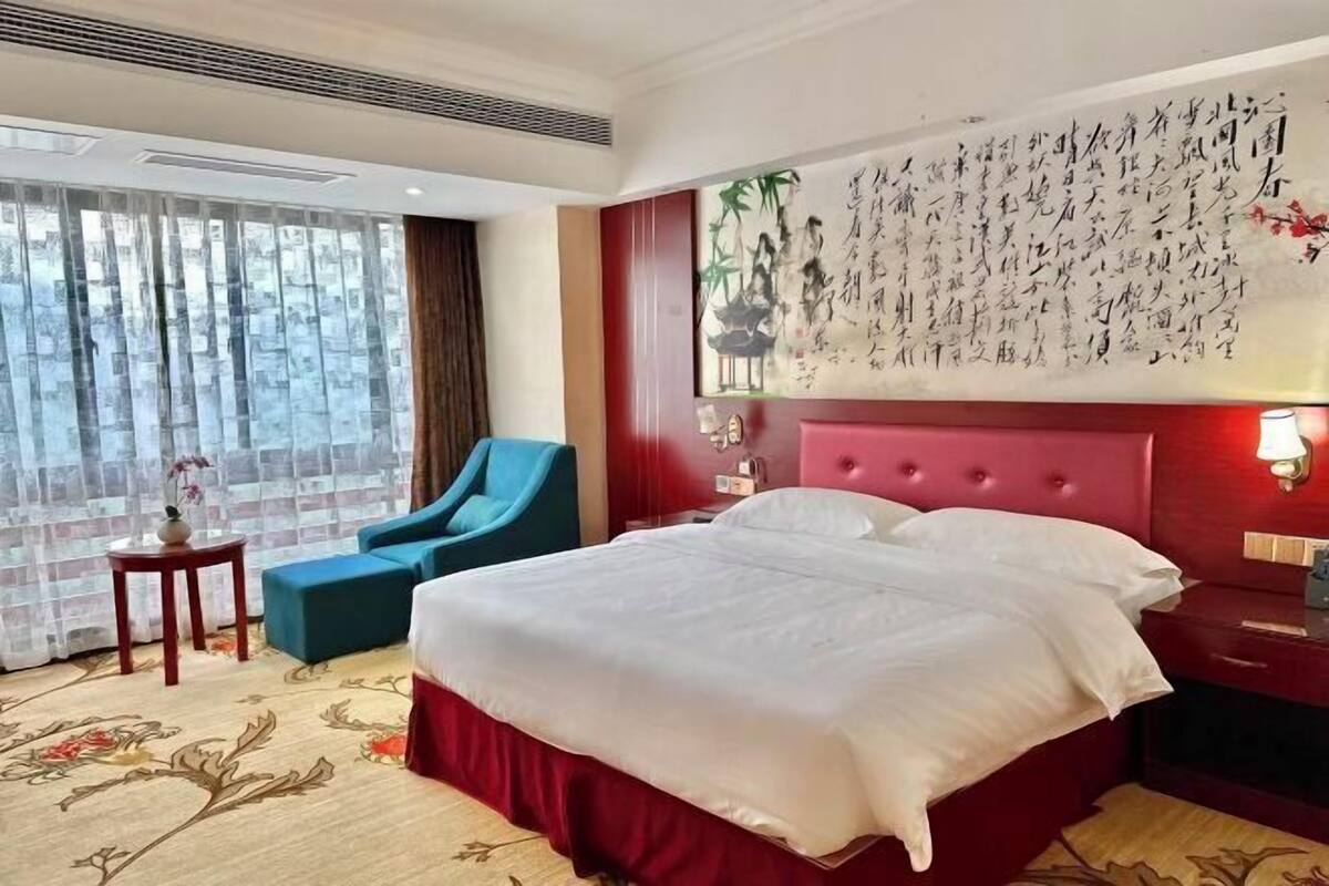 A hotel room with a large bed, a red headboard, a wall with calligraphy, a blue armchair, and a small table with a vase.