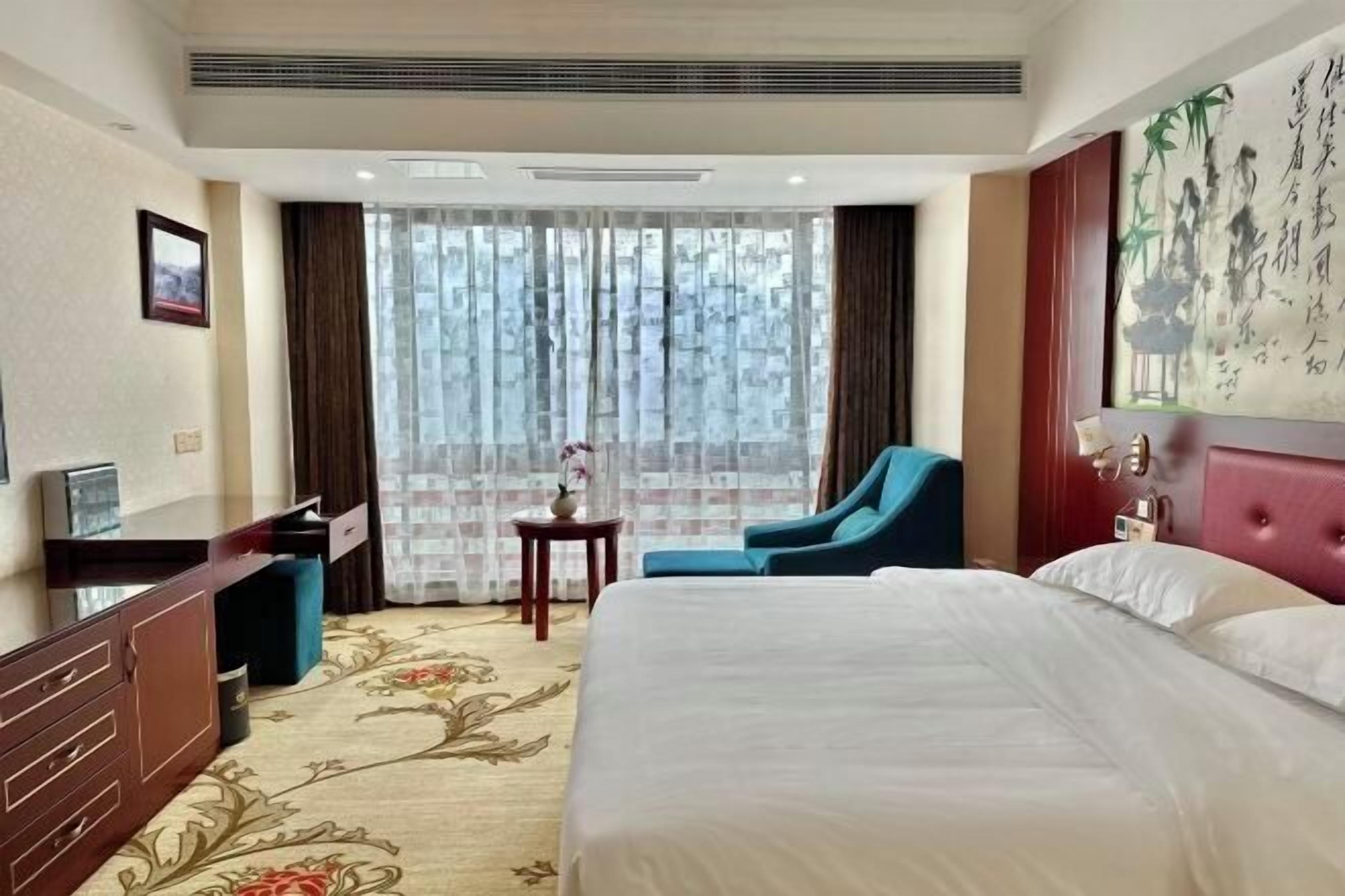 A hotel room with a large bed, a desk with a chair, a small table with a vase, and a large window with curtains.