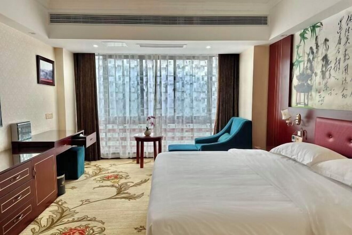 Habitación de hotel con una cama grande, un escritorio con silla, una mesita con un florero y un ventanal con cortinas.