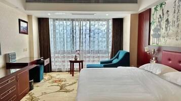 A hotel room with a large bed, a desk with a chair, a small table with a vase, and a large window with curtains.
