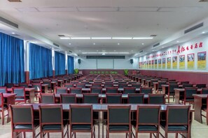 Un salón de conferencias con filas de mesas y sillas, un escenario al frente y carteles informativos en las paredes.