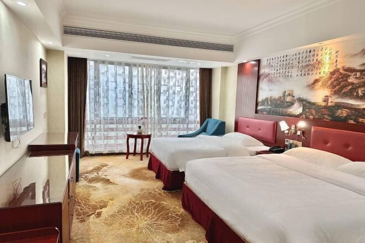 A hotel room with two beds, a TV, a painting on the wall, and a window with curtains.