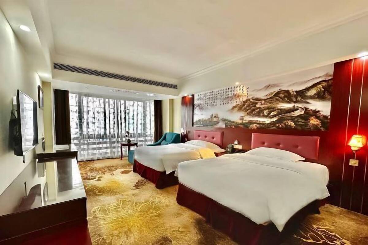 A hotel room with two beds, a large window, a mural, and a red accent wall.