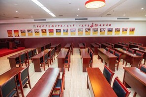 Une salle de conférence avec des rangées de tables et de chaises en bois, une moquette rouge et un mur orné d’affiches.