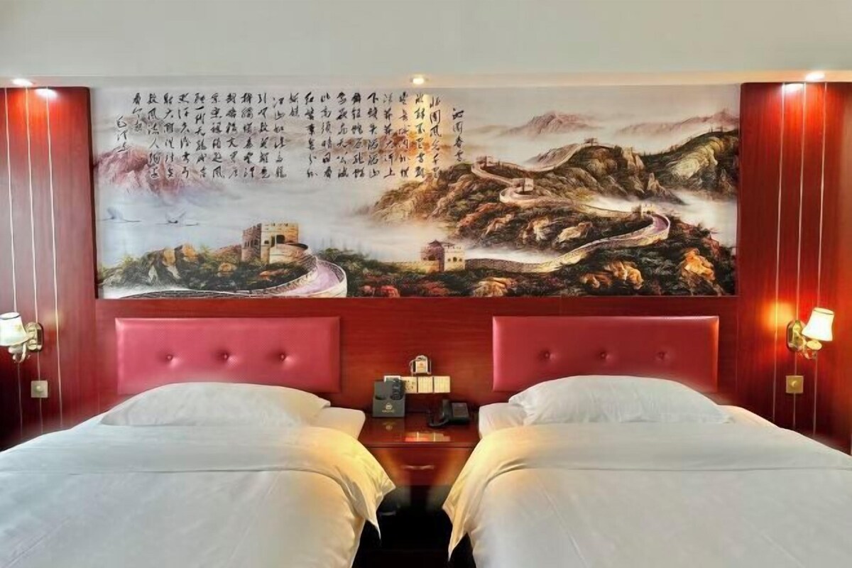 Habitación de hotel con un cuadro grande de un paisaje montañoso en la pared, un cabecero rojo y dos camas.