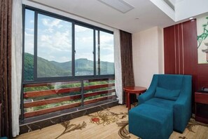 Una habitación con un ventanal amplio que ofrece vistas a colinas verdes, un sillón azul, una mesa redonda pequeña y un armario rojo.