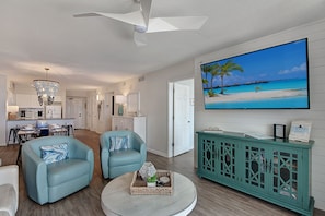 A living room with a large TV, teal furniture, and a kitchen in the background.