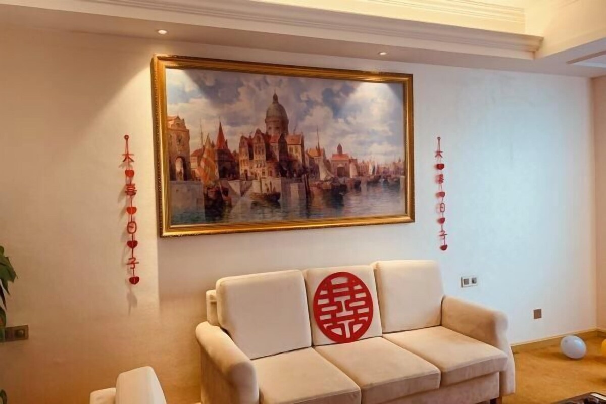 Ein Zimmer mit einem beigefarbenen Sofa, einem gerahmten Gemälde einer europäischen Stadtansicht und roten Dekorationselementen an den Wänden.