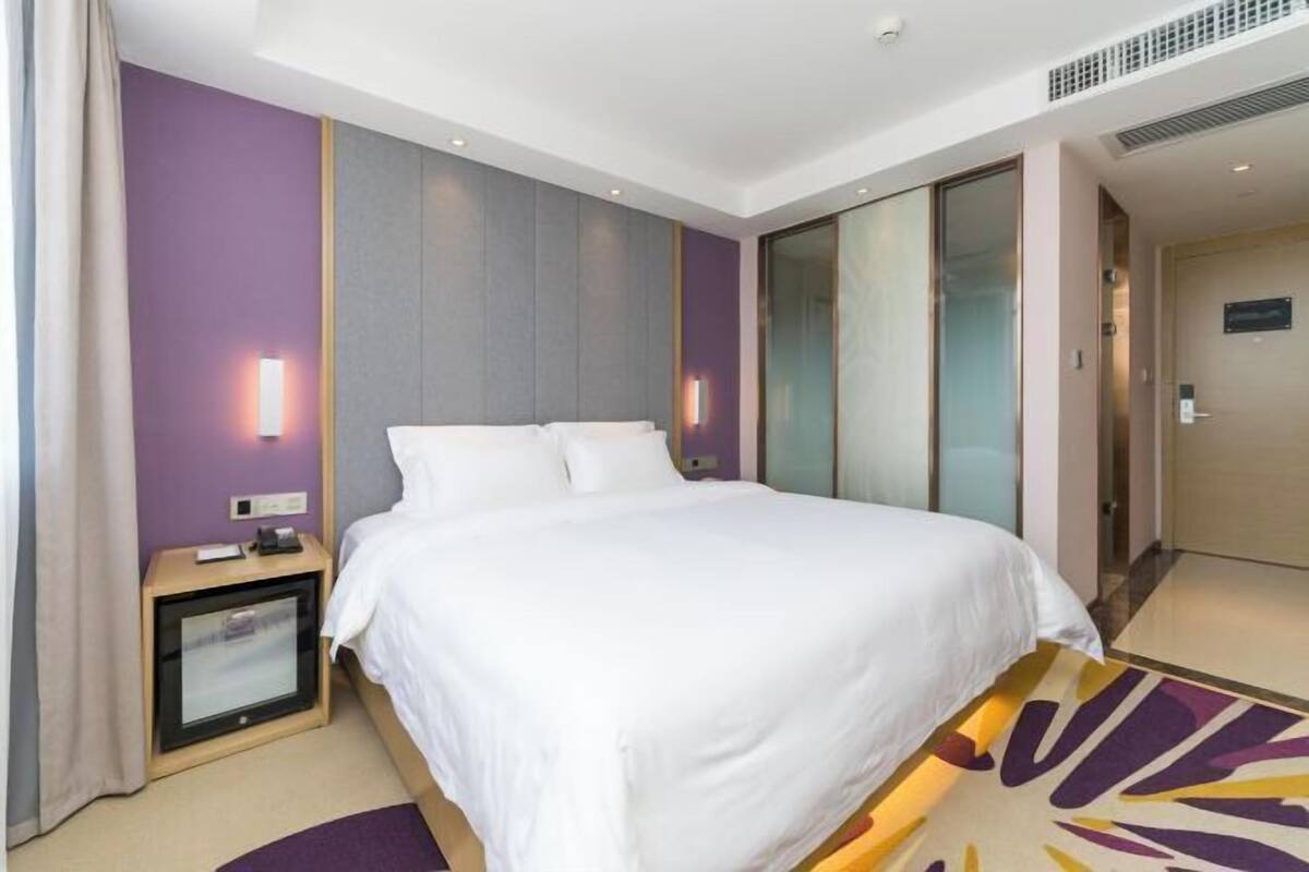 A hotel room with a large bed, a bedside table with a lamp, and a view of the hallway.