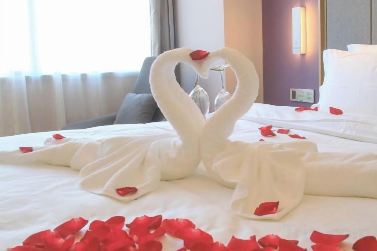 A bed with a towel arrangement in the shape of swans, a glass of water, and scattered rose petals.