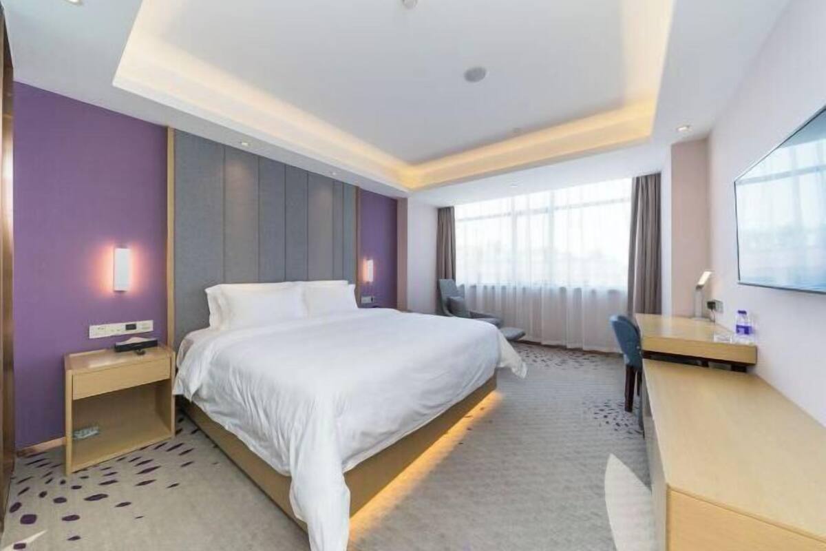 A hotel room with a large bed, a desk, and a chair. There is a window with curtains and a wall-mounted TV.