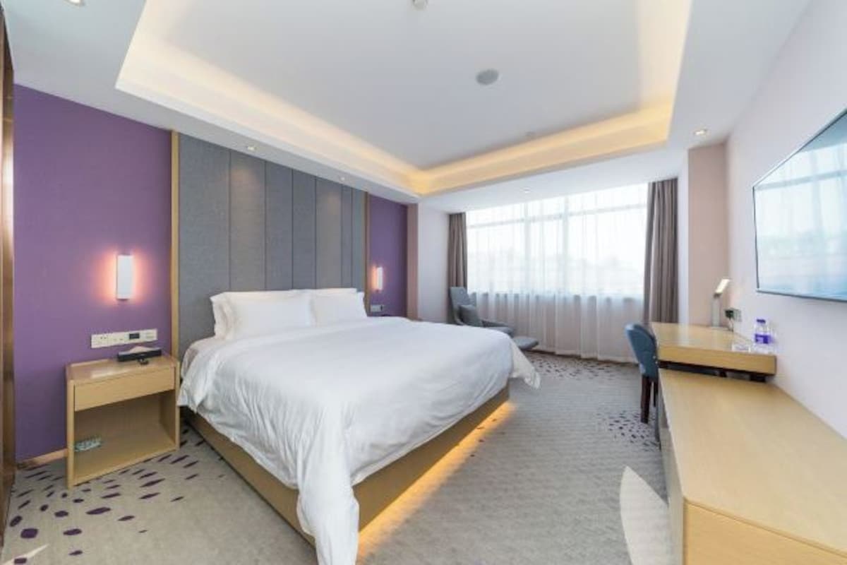 A hotel room with a large bed, a desk, and a chair. There is a window with curtains and a wall-mounted TV.