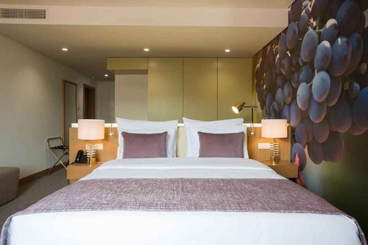 A hotel room with a large bed, two bedside tables with lamps, a wall-mounted artwork, and a door leading to another room.