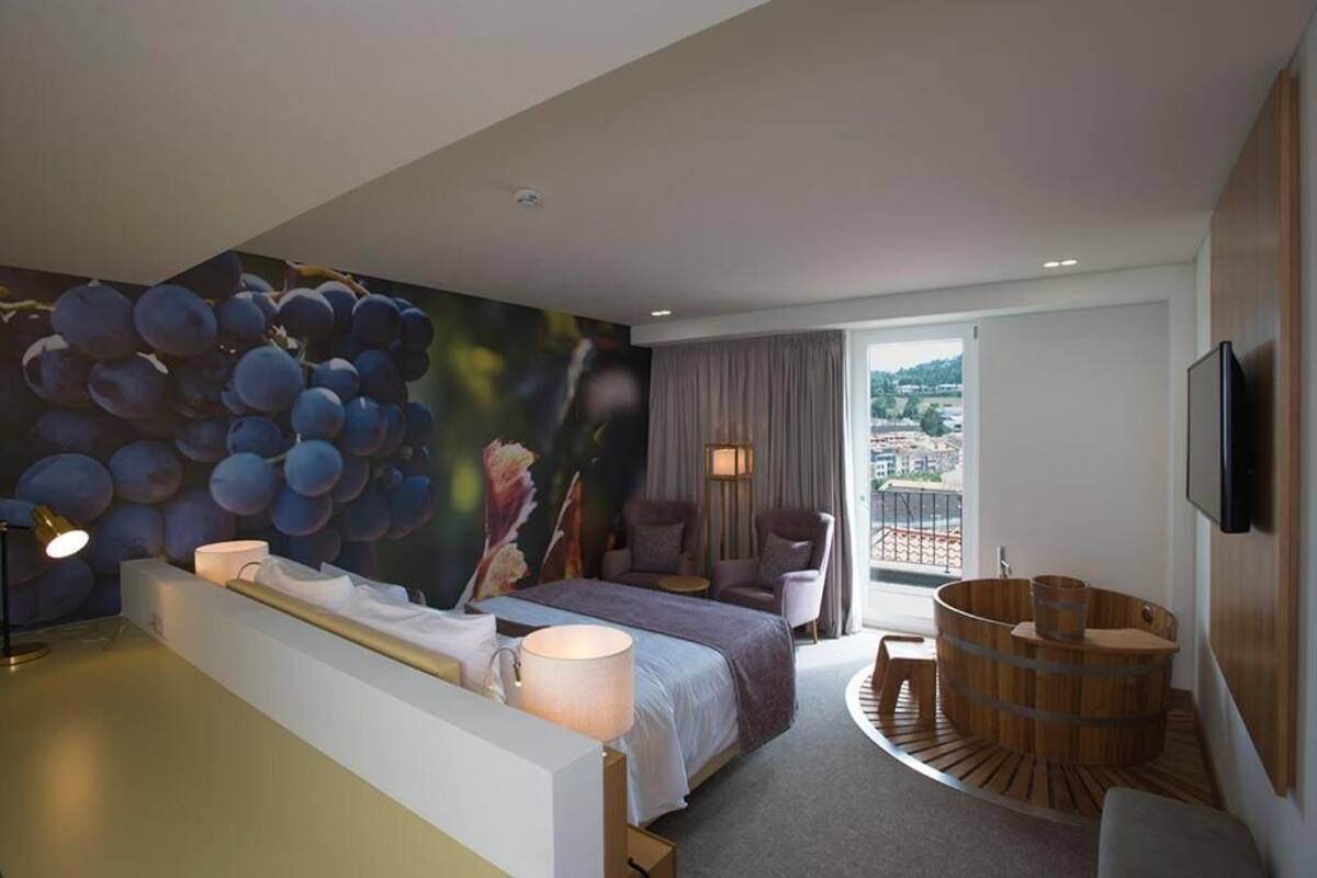 A modern hotel room with a large bed, a seating area with a view, a wooden bathtub, and a cityscape view through the window.