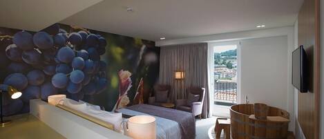 A modern hotel room with a large bed, a seating area with a view, a wooden bathtub, and a cityscape view through the window.